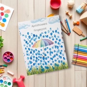 May include: A children's art project titled "April showers Fingerprint" featuring a colorful umbrella and blue fingerprint raindrops. The artwork is surrounded by art supplies, including crayons, paint, and a pink mallet, on a light wood surface.