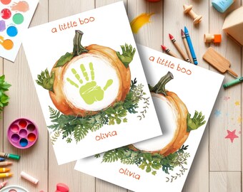 May include: Two watercolor prints featuring a pumpkin design with a green handprint in the center. The text "a little boo" is above the pumpkin, and the name "Olivia" is below. The pumpkins are surrounded by green foliage.