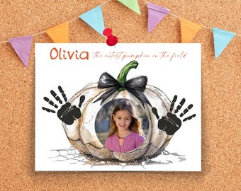 May include: A framed print featuring a watercolor pumpkin design with a black bow and green stem. The pumpkin has a circular cutout with a girl's photo. Black handprints are on either side. The text reads "Olivia the cutest pumpkin in the field."