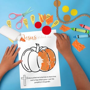 Puede incluir: Un proyecto de manualidades con un contorno de calabaza sobre papel blanco, parcialmente relleno de papel naranja. El texto "Jesus picked me" est&aacute; encima de la calabaza. Tijeras rosas, crayones y pegamento esparcidos sobre una superficie azul.