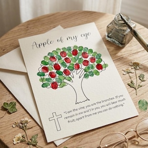 May include: A white greeting card featuring a black line drawing of a tree with red fingerprint apples and green fingerprint leaves. The text reads Apple of my eye and I am the vine; you are the branches. If you remain in me and I in you, you will bear much fruit; apart from me you can do nothing. A black cross is also printed.