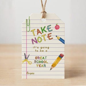 Puede incluir: Una etiqueta de regalo rectangular de papel con un patrón de papel de cuaderno rayado y un lazo de cuerda de yute. El texto dice TAKE NOTE it's going to be a GREAT SCHOOL YEAR from. Las ilustraciones incluyen un clip verde, una manzana roja, un lápiz azul, unas tijeras amarillas y un lápiz amarillo.