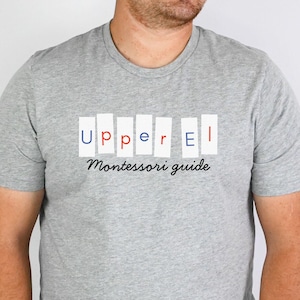 May include: A gray t-shirt with the text "Upper El Montessori guide" in a rectangular design. The word "Upper" is in blue and red, and "El" is in white. The shirt is short-sleeved and appears to be made of a soft material.