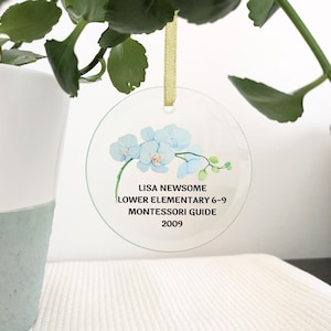 May include: A round, clear glass ornament with a blue orchid design. The ornament has the text "Lisa Newsome Lower Elementary 6-9 Montessori Guide 2009" printed on it.