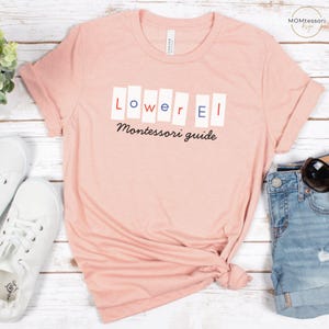May include: A light pink t-shirt with the text "Lower El Montessori guide" in a mix of red, blue, and black. The shirt is folded at the bottom and is paired with denim shorts and white sneakers.