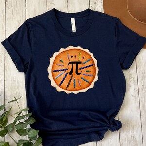 May include: Navy blue t-shirt featuring a pie graphic with the mathematical symbol pi in the center. The pie design includes colorful beads and a crust. The shirt is made of a soft material and is suitable for casual wear.