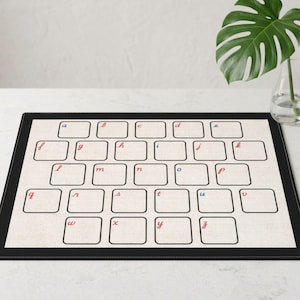 May include: A rectangular placemat with a black border and a light beige textured background. It features a grid of twenty-six empty rounded squares, each labeled with a lowercase letter of the alphabet in cursive script, alternating between red and blue.