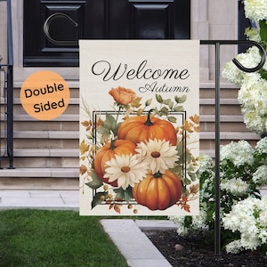Double-sided garden flag with a fall theme. The flag features the words "Welcome Autumn" above a design of orange pumpkins, white daisies, and fall foliage. The flag is displayed on a black metal stand in a garden setting.
