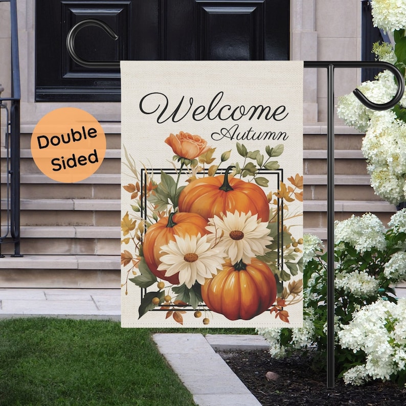 Double-sided garden flag with a fall theme. The flag features the words "Welcome Autumn" above a design of orange pumpkins, white daisies, and fall foliage. The flag is displayed on a black metal stand in a garden setting.