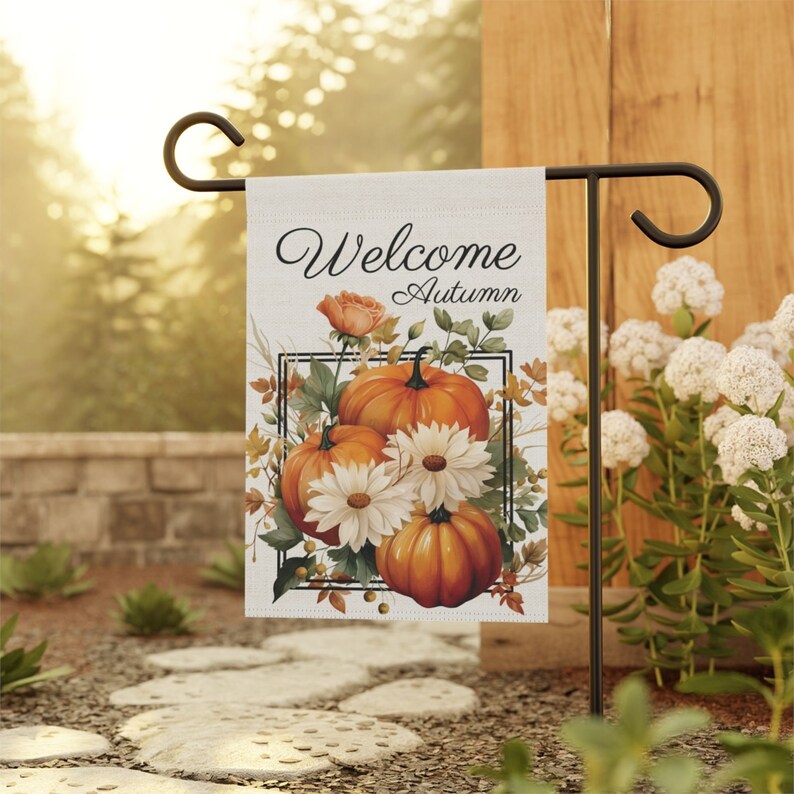 A rectangular garden flag with the words "Welcome Autumn" in script. The flag features a watercolor design of orange pumpkins, white daisies, and fall foliage within a black rectangular frame.