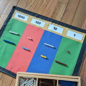 May include: A Montessori math learning tool with a colorful mat and wooden beads. The mat is divided into green, red, and blue sections, labeled 1000, 100, 10, and 1. Various colored beads are arranged on the mat, with a wooden box of beads in the foreground.