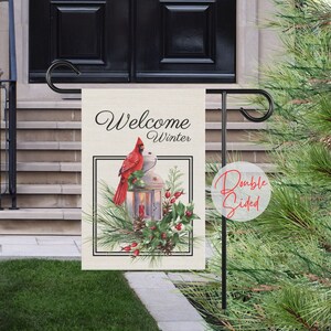 May include: A double-sided garden flag with the words "Welcome Winter" in script. The flag features a red cardinal perched on a lantern with a lit candle, surrounded by pine needles and berries. The flag is displayed on a black metal stand.
