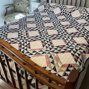 May include: A bed quilt featuring a repeating geometric pattern of cream, navy blue, and dusty pink fabric squares. The design consists of large central cream blocks surrounded by intricate navy and pink triangular star-like motifs.