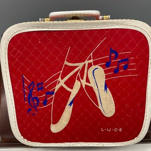 May include: Red, quilted suitcase with a white trim and handle. The front features a ballet-themed design with ballet shoes, musical notes, and the word "L.U.C.E." in white. The suitcase has a rectangular shape.