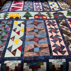 May include: A colorful quilt with a patchwork design featuring a schoolhouse, trees, and a school bus. The quilt is made with various fabrics in shades of blue, pink, green, yellow, and red.