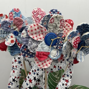 May include: A bouquet of fabric flowers with petals in various patterns and colors, including red, white, and blue. The flowers are made from quilted fabric and have blue button centers. The stems are wooden dowels wrapped in star-patterned fabric.