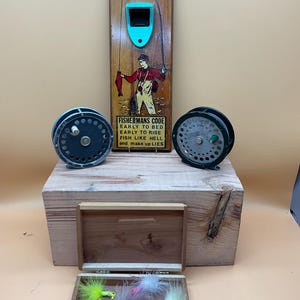 Può includere: Un apribottiglie da parete in legno a tema pesca con un apribottiglie turchese e una grafica di un pescatore. Sono visibili anche due mulinelli da pesca vintage e una scatola di legno con mosche da pesca. Il testo sull'apribottiglie recita "FISHERMANS CODE".