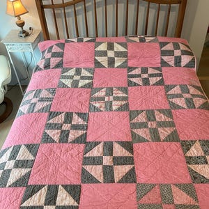 May include: A handmade quilt with a pink background and geometric patterns in gray, white, and red. The quilt is spread across a wooden bed frame. The quilt's design features squares and triangles.