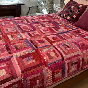 Op de afbeelding: Een patchwork quilt met een log cabin patroon in diverse tinten roze, magenta en rood. De quilt is samengesteld uit talloze stoffen vierkanten met uiteenlopende texturen en patronen, waaronder bloemmotieven, stippen en effen stoffen.