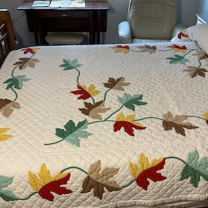 May include: A white quilted bedspread featuring a leaf design. The leaves are in shades of red, yellow, brown, and green, connected by green stems. The bedspread is on a bed with a wooden frame.
