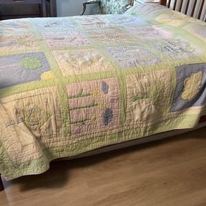 May include: A patchwork quilt featuring a grid of squares with embroidered nature-themed designs including trees, flowers, and leaves in soft pastel shades of green, yellow, blue, and pink, bordered by a light green gingham fabric trim.