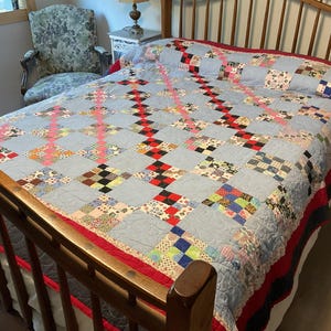 May include: A handmade quilt with a light blue background and a patchwork design. The quilt features squares of various colors and patterns, including red, black, and floral prints. The quilt is on a wooden bed frame with a red border.