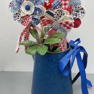 May include: A bouquet of fabric flowers in a blue speckled pitcher. The flowers are made from various patterned fabrics in red, white, and blue. A blue ribbon is tied around the pitcher's handle. Green leaves add a natural touch.