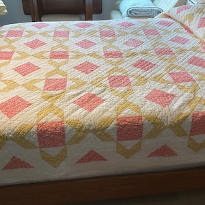 May include: A handmade quilt with a repeating pattern of pink and yellow diamonds on a white background. The quilt is on a bed with a wooden frame. The quilt's design includes pink squares and yellow diamond shapes.