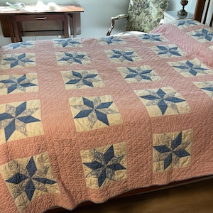 May include: A handmade quilt with a pink background and a repeating pattern of blue and cream-colored starbursts. The quilt is spread over a wooden bed frame, showcasing its intricate stitching and design. The quilt is a cozy and decorative bedding item.