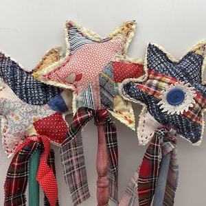 May include: Three star-shaped fabric ornaments attached to painted wooden sticks. Each star features a patchwork design of various patterned textiles in red, blue, and white. The sticks are adorned with tied fabric ribbons in plaid and polka dot patterns.