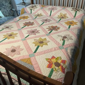 May include: A handmade quilt with a floral pattern. The quilt features large, colorful flower appliqués in shades of yellow, pink, and brown, set against a white background. Green stems connect the flowers. The quilt is on a wooden bed frame.
