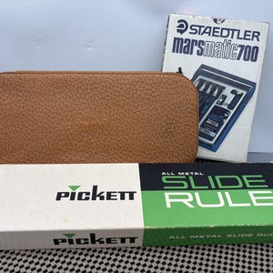 May include: A brown leather case, a box labeled "STAEDTLER marsmatic 700", and a box with "PICKETT" and "ALL METAL SLIDE RULE" printed on it. The boxes are white, green, and black.