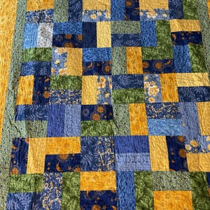 May include: A handmade quilt with a patchwork design. The quilt features squares of various fabrics in shades of blue, green, and yellow. Some squares have floral patterns, while others have solid colors or textured designs. The quilt is bordered with a yellow floral fabric.
