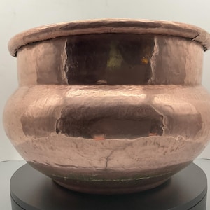 May include: A hammered copper pot with a wide, rounded body and a rolled rim. The pot has a warm, reddish-brown color and a textured surface. The pot is likely used for decorative purposes.