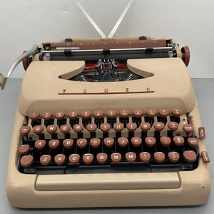 May include: A beige Tower President typewriter featuring a QWERTY keyboard with round, dark brown keys. The machine includes a metal carriage return lever on the left, a paper bail, and a visible ruler along the top of the platen area.