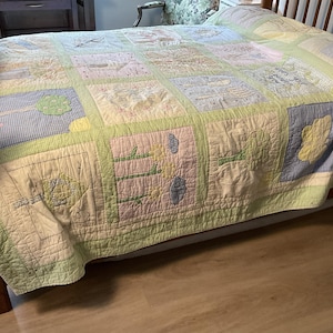 Puede incluir: Un edredón de retazos con un borde verde claro y cuadrados en colores pastel. Los cuadrados presentan diseños bordados de árboles, nubes y otros elementos temáticos de la naturaleza. El edredón está sobre una estructura de cama de madera.