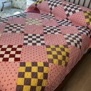 May include: A patchwork quilt featuring a repeating pattern of solid pink floral fabric squares alternating with multi-colored checkered blocks. The checkered squares consist of various combinations of dark red, yellow, and dark brown small-scale prints.