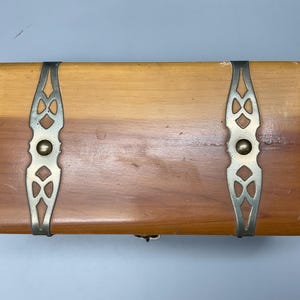 May include: Rectangular wooden box with a light brown finish, accented with decorative silver-toned metal brackets and a small clasp. The box has a natural wood grain and a simple, classic design.