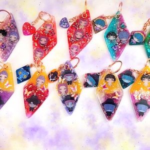 May include: A collection of handmade keychains with diamond-shaped resin pendants. Each keychain features a miniature character design, glitter, and a small charm. The keychains are in various colors, including pink, purple, blue, and yellow.