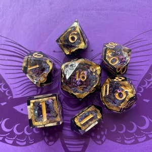 Può includere: Set di sette dadi viola e dorati con glitter iridescente incorporato nella resina. I dadi sono numerati da 1 a 6 e 16. I dadi sono su uno sfondo viola con un motivo a farfalla.