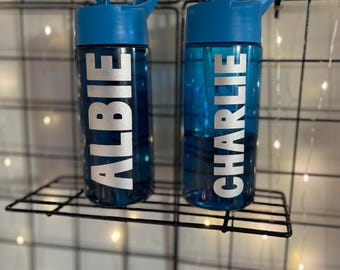 Può includere: Due borracce blu con tappi blu e scritte bianche "ALBIE" e "CHARLIE". Sono esposte su una mensola di filo nero su uno sfondo a griglia con luci a catena.