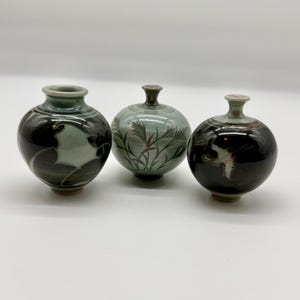 May include: Three small, round ceramic vases with lids. The vases are painted in shades of black, green, and gray, with floral and abstract designs. The vases are displayed on a white surface.