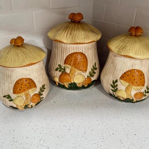 May include: Three ceramic mushroom-shaped canisters with lids. The canisters are off-white with a textured, vertical pattern. Each canister features a large, orange mushroom with brown spots and smaller mushrooms. The lids are light yellow with a textured, ribbed design.