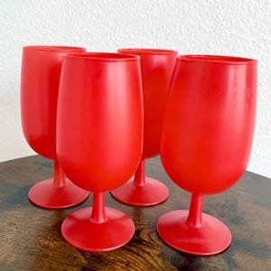 May include: A set of four matching red plastic goblets with a smooth, matte finish. Each goblet features a rounded bowl, a slender stem, and a circular base, standing upright in a group arrangement to display their uniform vibrant color.