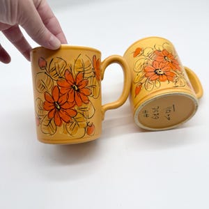 May include: Two yellow ceramic mugs with orange floral designs. The mugs have curved handles and are decorated with orange flowers and green leaves. The mugs are on a white surface.