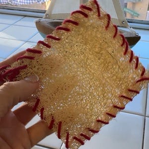 Puede incluir: Una esponja de lufa cuadrada beige con costuras rojas en los bordes. La lufa se sostiene en una mano y un pequeño velero de madera es visible en el fondo.