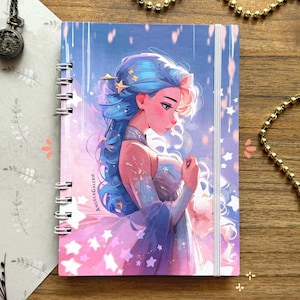 Könnte beinhalten: Spiralgebundenes Notizbuch mit einer Fantasy-Illustration einer Frau mit blauen Haaren und einem sternenbesetzten Kleid. Das Cover hat ein rosa-blaues Farbschema mit dem Text "AngelaGalerie". Das Notizbuch liegt auf einer Holzoberfläche.