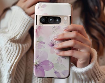 May include: A phone case with a floral design featuring purple and pink flowers on a white background. The case has a rectangular shape with a camera cutout and a gold-colored frame. The case is held in a person's hands.