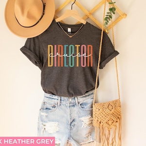 May include: Dark heather grey V-neck t-shirt with the text "cruise director" in rainbow colored letters. The shirt is being modeled on a person wearing denim shorts and a straw hat.