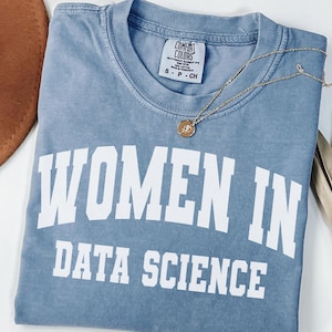 May include: A blue short-sleeve t-shirt with a faded wash finish. The shirt features the white text "WOMEN IN DATA SCIENCE" printed in a bold, arched collegiate font across the chest. A small gold-toned pendant necklace rests on the neckline.